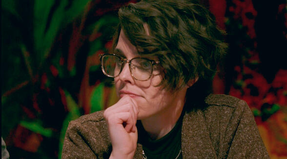 Mellie Doucette is pictured. She has short wavy brown hair in an asymmetrical cut, white skin, and large glasses. She sits with her fist on her chin in thought.