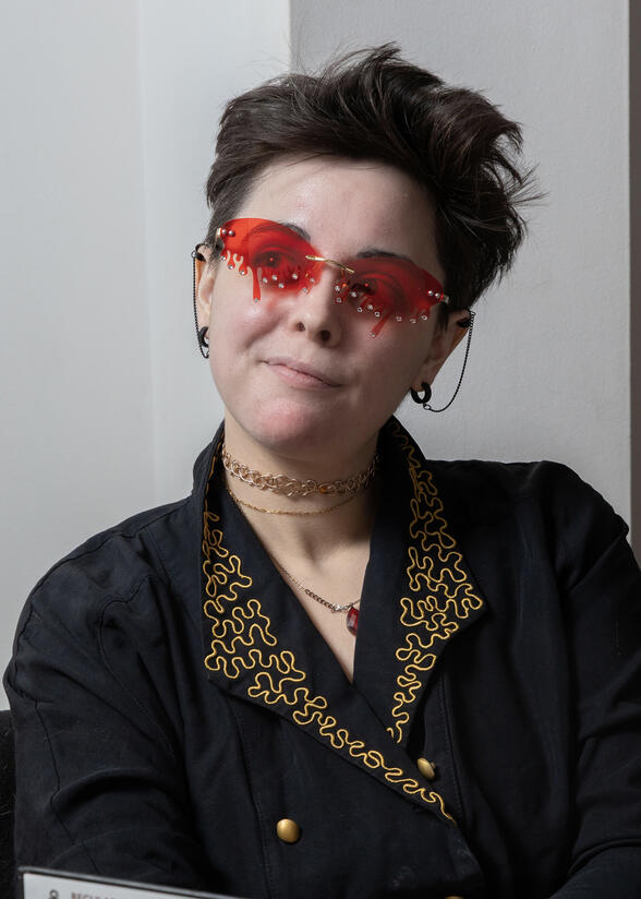 Mellie Doucette is pictured. She has short wavy brown hair in an asymmetrical cut, white skin, and large glasses. She sits with her fist on her chin in thought.