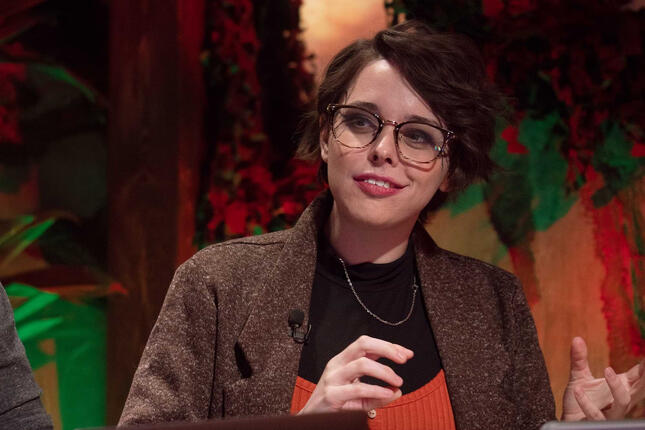 Mellie Doucette is pictured. She has short wavy brown hair in an asymmetrical cut, white skin, and large glasses. She sits with her fist on her chin in thought.